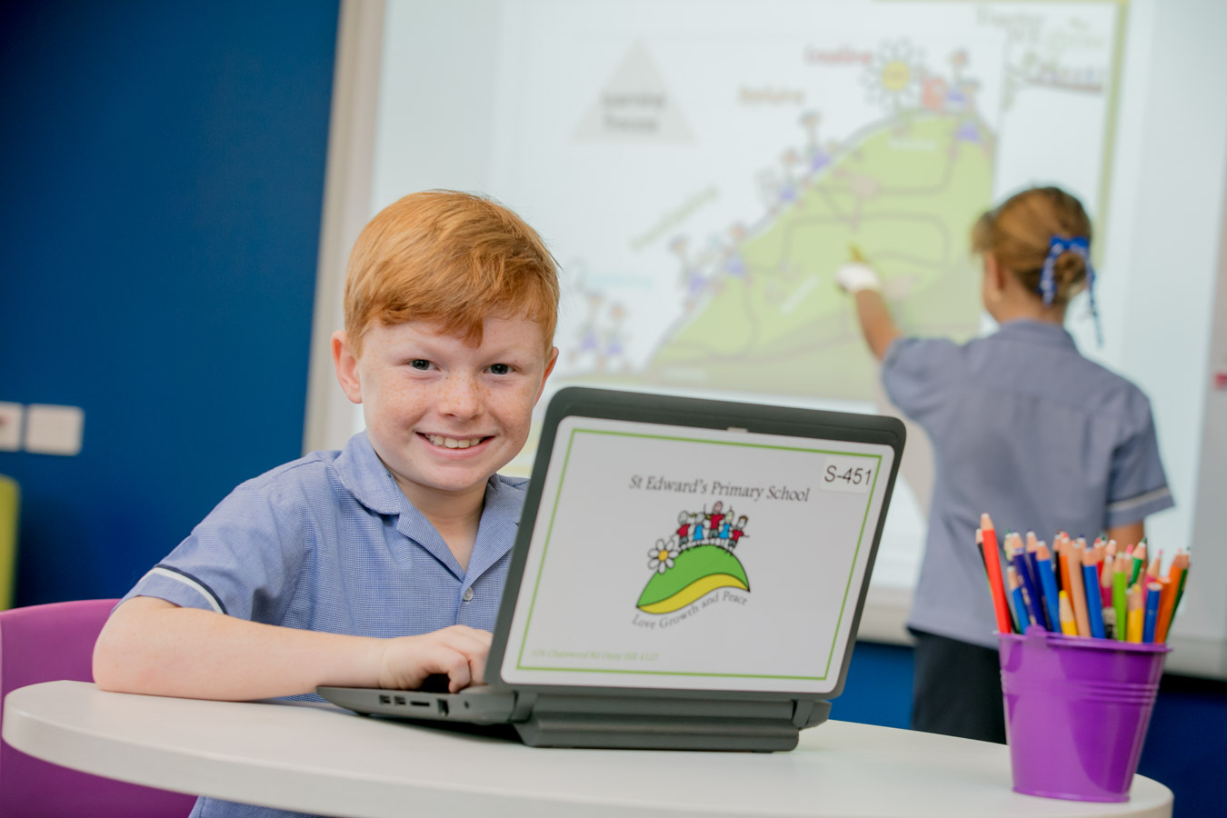 Student using a laptop in a classroom while a classmate interacts with a digital display highlighting engagement in technology supported learning and collaboration